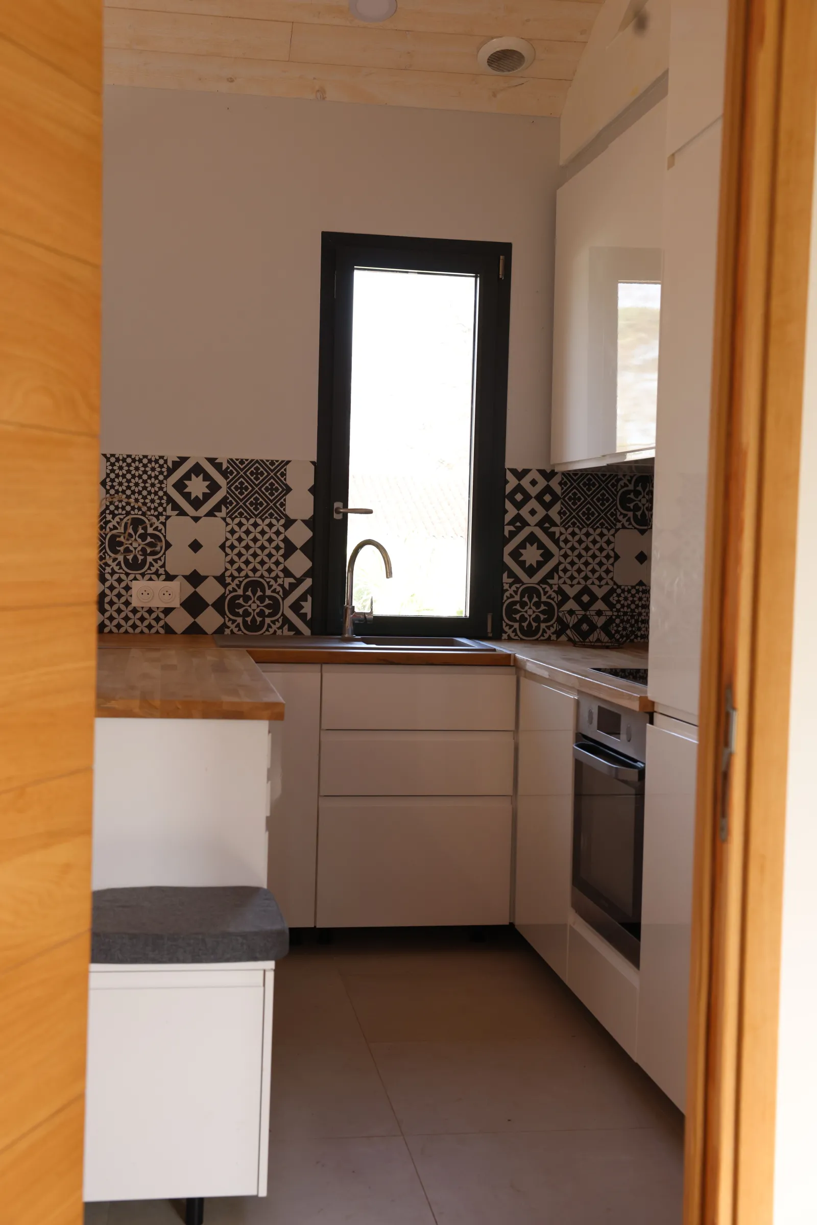 Modern kitchen in an eco-cottage featuring black and white geometric patterned tiles, black-framed window, gray countertops, beige cabinets, and yellow door on the left