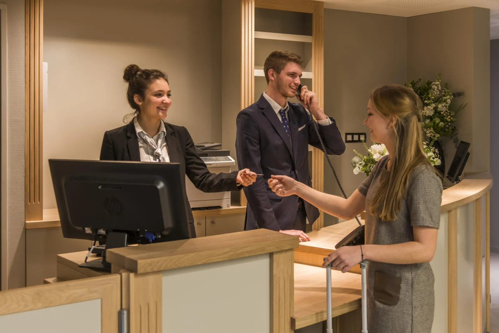 Three professionals at the reception desk of Hôtel Vatel in Bordeaux: a woman in a black blazer behind the counter, a man in a navy suit on the phone, and a blonde female guest holding a folder.