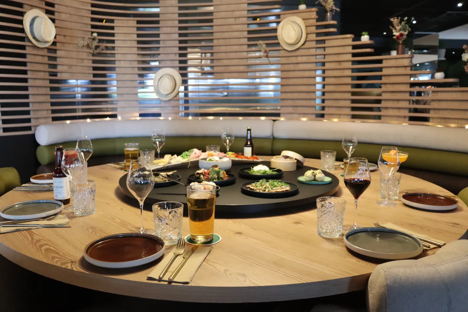 Les Décantés restaurant dining room in Bordeaux featuring a round wooden table set with elegant place settings, wine glasses, rotating tray of gourmet dishes, green banquette seating, and wooden wall with circular windows.