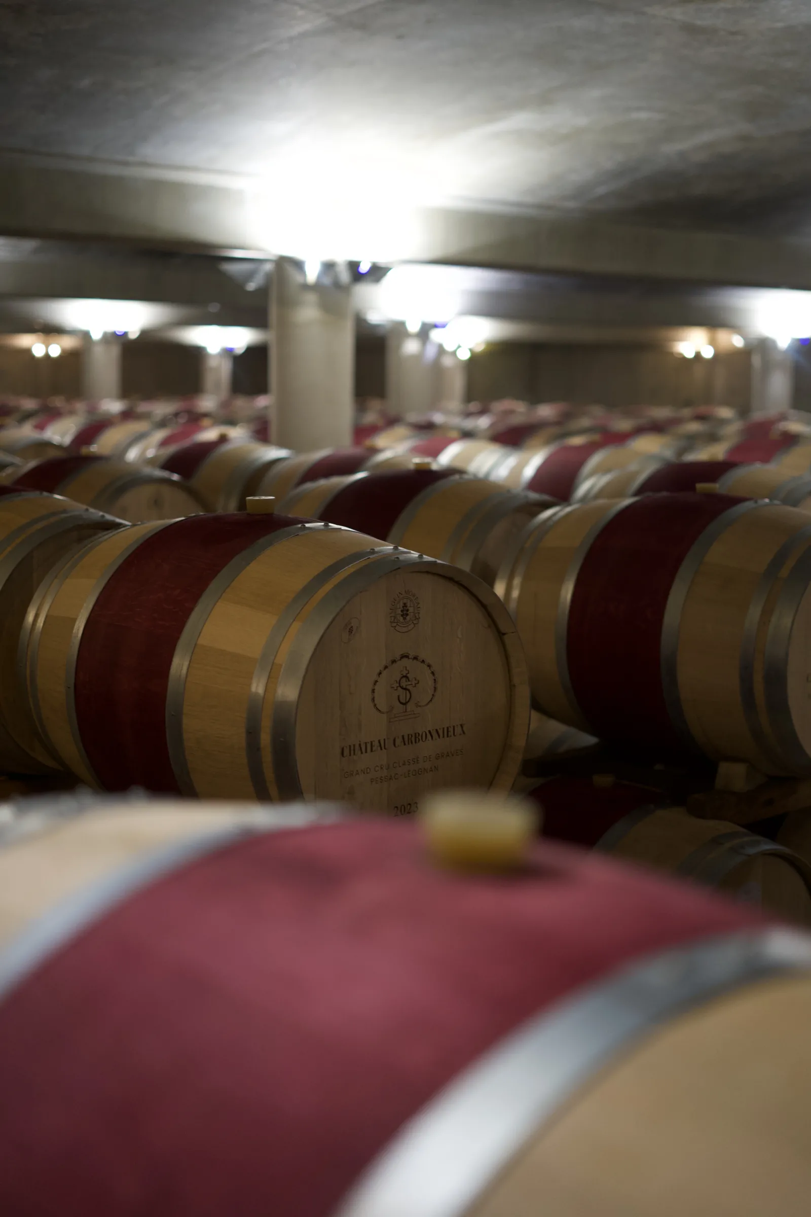 Wine cellar at Château Carbonnieux in Bordeaux featuring rows of oak barrels with red and golden bands under interior lighting.