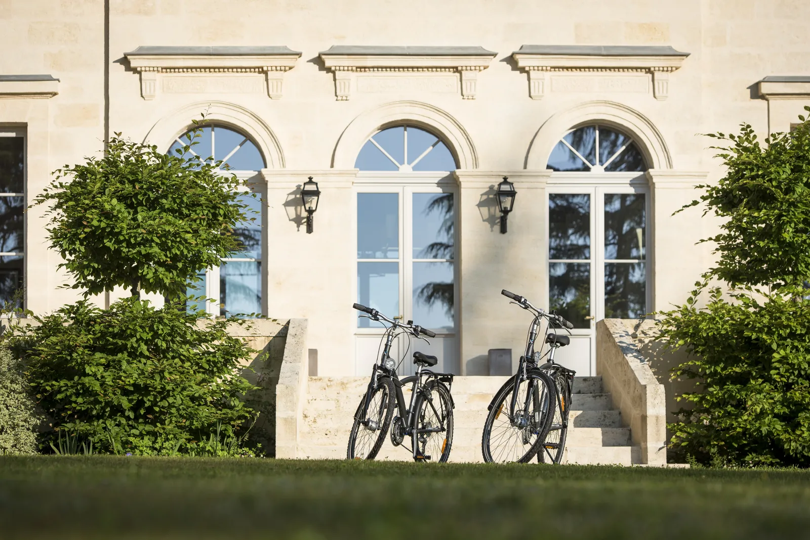 Two bicycles parked in front of the classical facade of Chartreuse Haut-Bailly in Bordeaux, featuring three large arched windows, surrounded by greenery and stone steps.