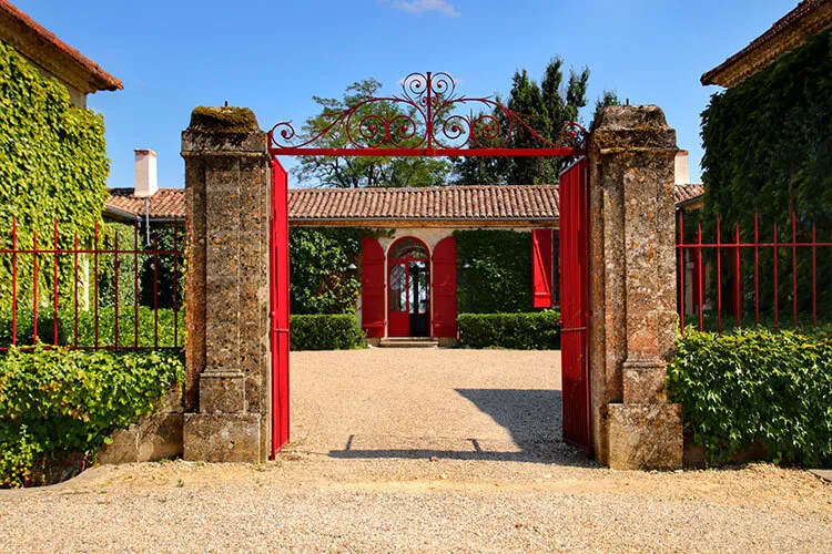 Red entrance gate of Château Sigalas Rabaud in Bordeaux, framed by stone columns with manicured gardens and ivy, leading to a paved courtyard