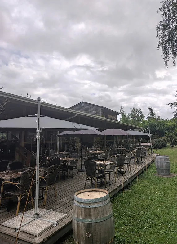 Outdoor terrace of Bistrot du Lac in Bordeaux featuring dining tables and chairs under white umbrellas, wine barrels as decoration, and green lawn in the background.