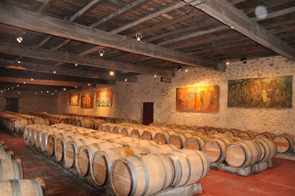 Wine cellar at Château Sigalas Rabaud in Bordeaux featuring rows of wooden barrels lined up on terracotta flooring, white stone walls, and exposed wooden beams