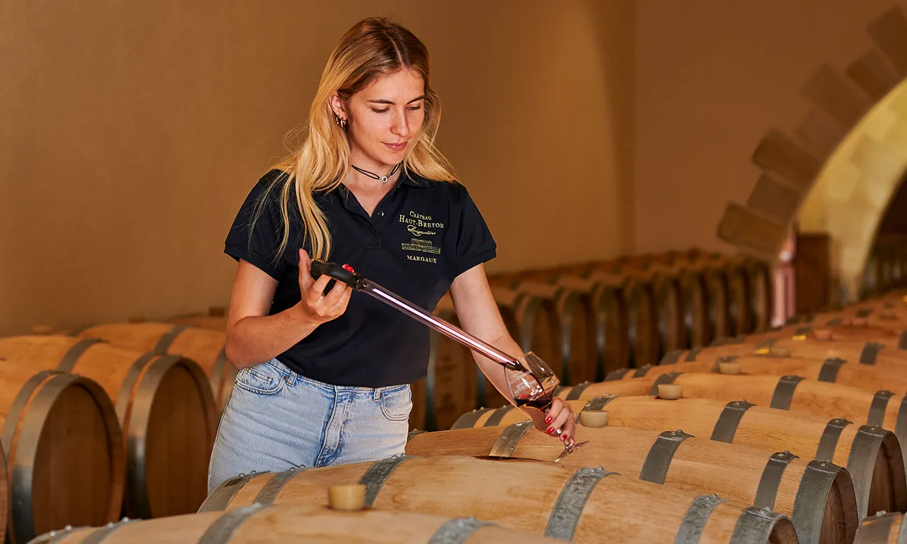 A woman in a black Château Haut Breton polo shirt samples red wine from an oak barrel in the winery cellar in Bordeaux.