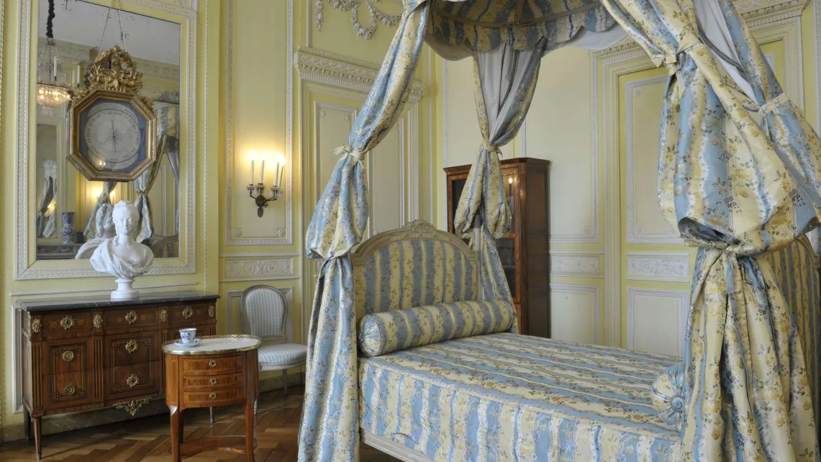 Eighteenth-century bedroom at the Bordeaux Museum of Decorative Arts, featuring a four-poster canopy bed draped with blue and gold fabrics, pale yellow ornate walls, gilt mirror, inlaid wooden dresser, and period furniture.