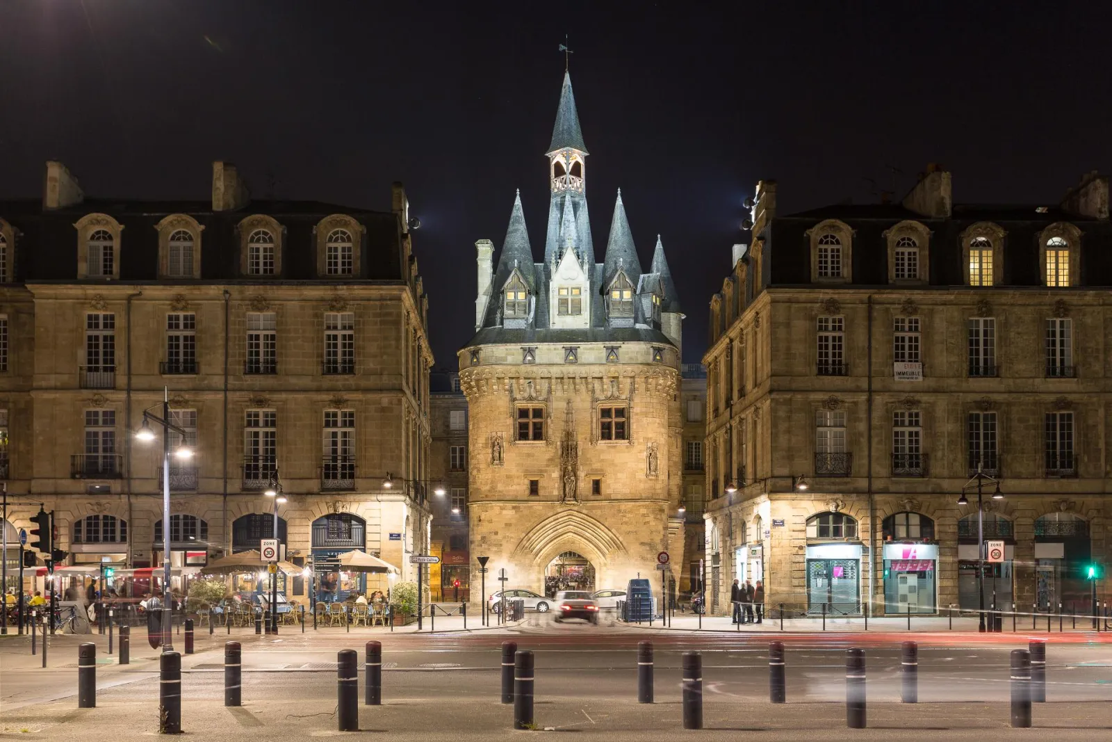 Porte Cailhau lit up at night in Bordeaux, medieval Gothic tower with pointed spires surrounded by Haussmann-style buildings, paved square with bollards in foreground.
