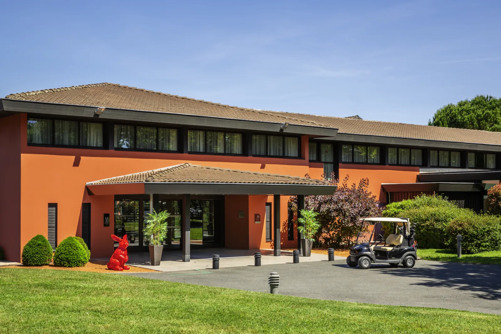 Modern façade of Cabot Bordeaux MGallery Collection hotel featuring orange and red walls, main entrance, golf course with cart, landscaped grounds and clear blue sky.