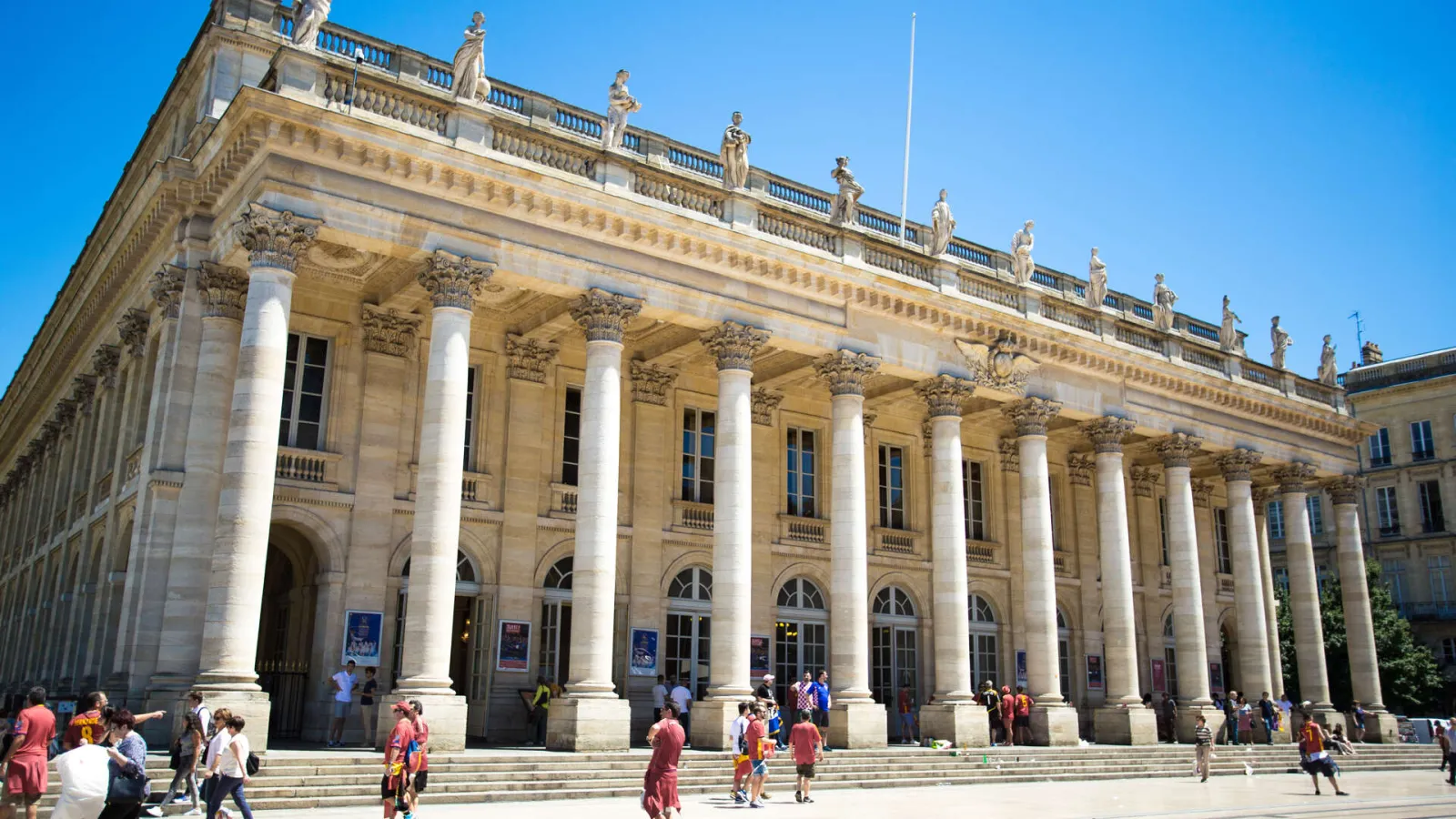 The Grand Theatre of Bordeaux featuring its neoclassical stone facade, white Corinthian columns, and decorative statues, set against a clear blue sky with visitors in the foreground.