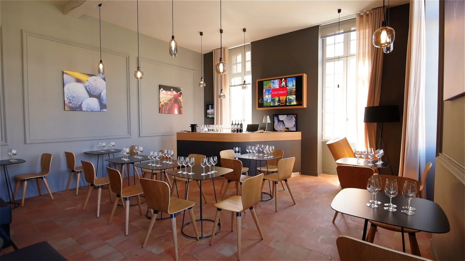 Modern tasting room at the Maison du Vin de Saint-Emilion in Bordeaux, featuring black tables, light wood chairs, wine glasses, wooden and black bar counter, digital displays, hanging pendant lights, and gray and brown walls.