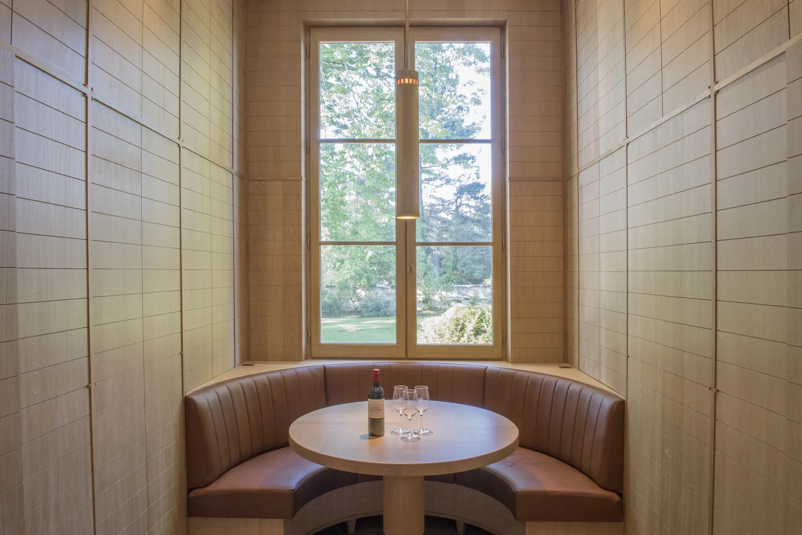 Intimate dining nook with brown leather banquette seating, round white table, wine bottle and glasses, framed by large windows overlooking the Chartreuse Haut-Bailly vineyards near Bordeaux