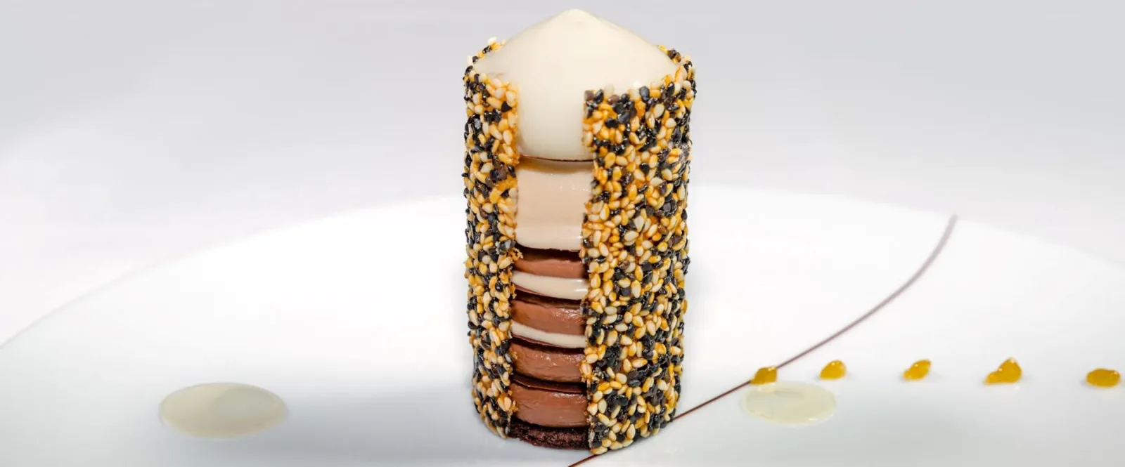Saint-James Bordeaux stacked dessert: alternating layers of chocolate and vanilla cream, coated with black and golden sesame seeds, topped with a white cream sphere, plated on white with sauce line and fruit pearls.