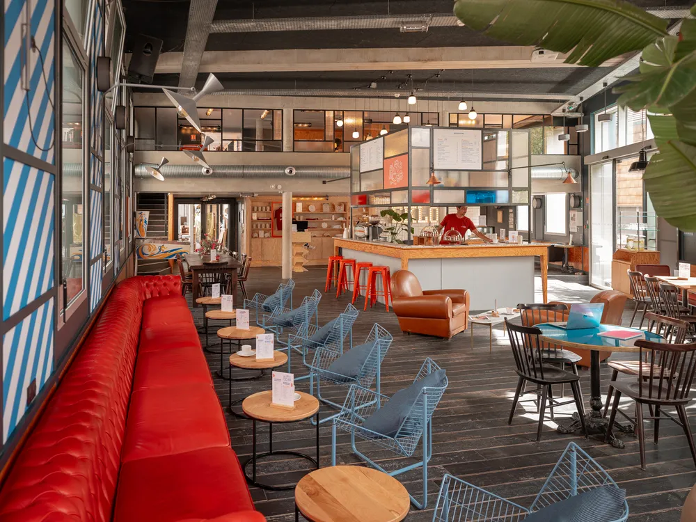 Modern common area at Eklo Bordeaux featuring a large red sofa, blue chairs, wooden high tables, a kitchen counter, and bright welcoming atmosphere.