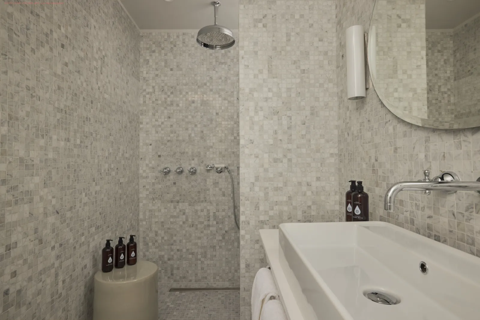 Modern bathroom with walk-in shower, light beige mosaic tiles, white bathtub, chrome showerhead and brown toiletry bottles on display.