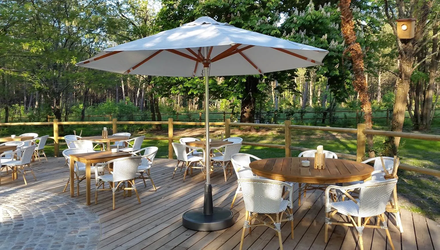 Outdoor terrace at Château Léognan in Bordeaux featuring round and square tables, white wicker chairs, blue and white umbrella, surrounded by lush forest and wooden fencing
