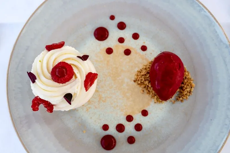 Gourmet dessert plate featuring whipped cream decorated with raspberries, red coulis and biscuit crumbs, presented on a white base with decorative coulis pearls