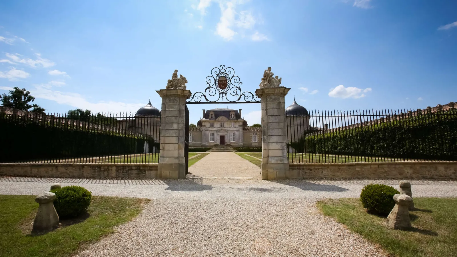 Overview of Château de Malle in Bordeaux, featuring ornate stone gateways with sculptures, wrought iron fencing, gravel drive, and a classical mansion with mansard roof.