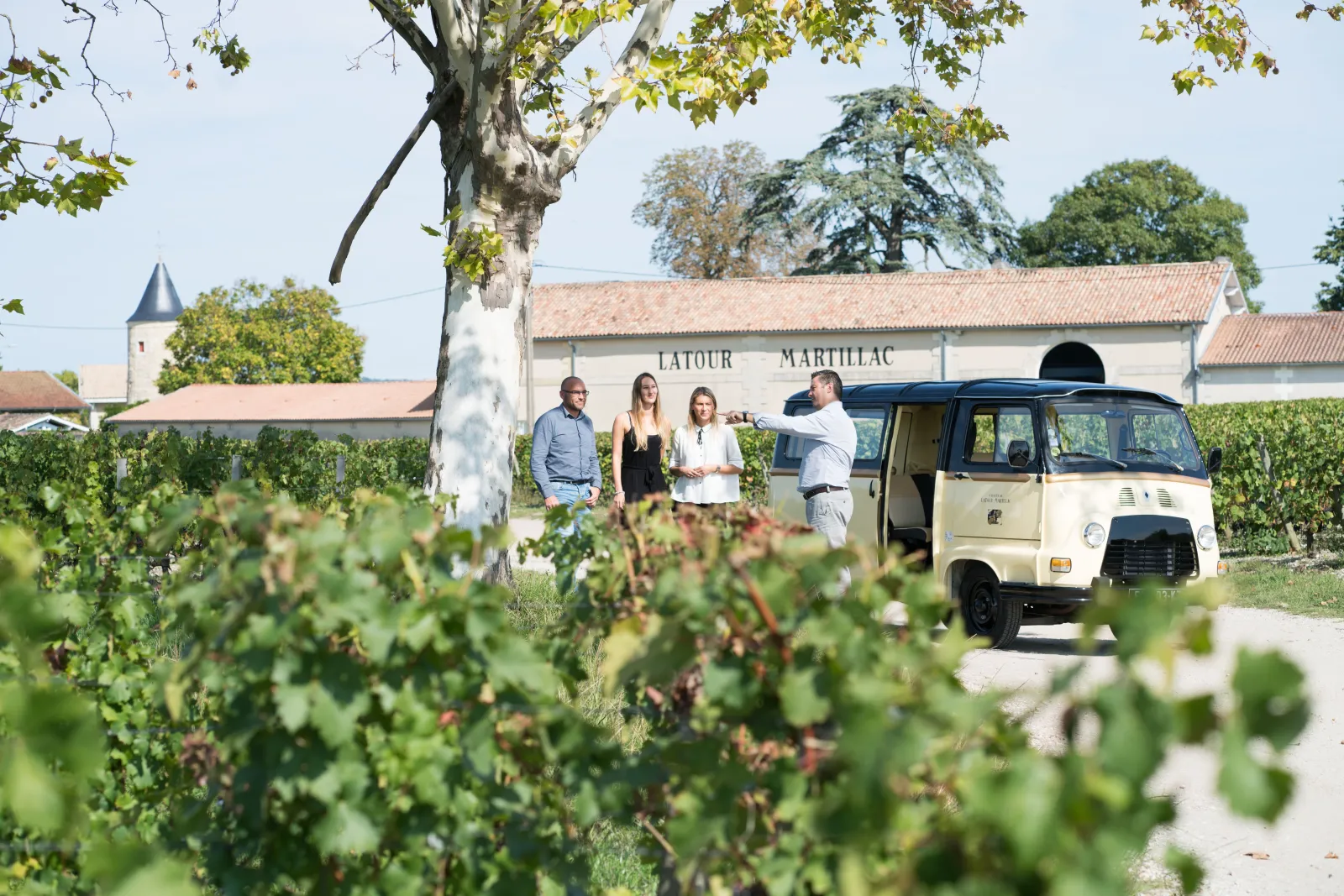 Guided tour at Château Latour-Martillac in Bordeaux with visitors gathered near a vintage tour vehicle, surrounded by vineyards and the historic winery building.