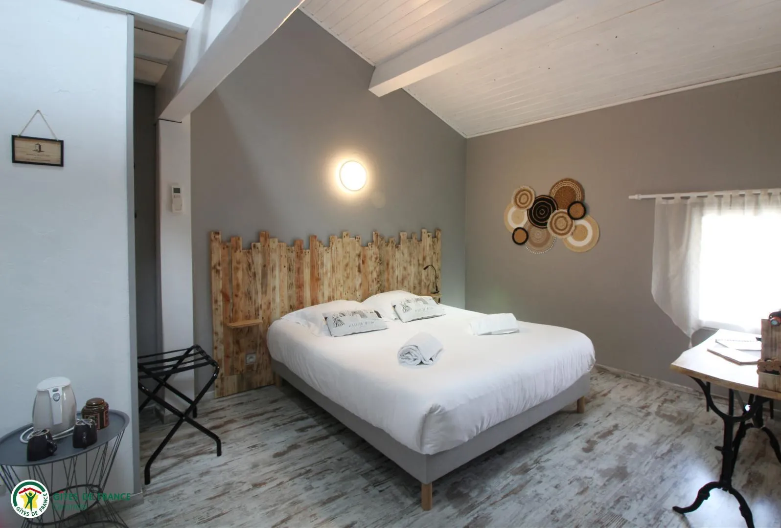 Modern guest room at Château Pierre de Montignac in Bordeaux, featuring a white double bed with wooden headboard, decorative woven baskets on the wall, white curtains, and contemporary furnishings.