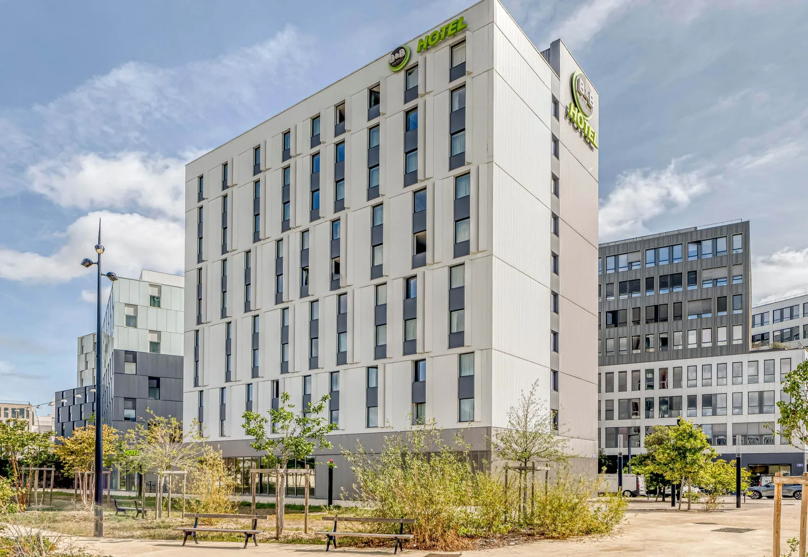 Facade of B&B Hotel Bordeaux Centre Gare Saint-Jean, a modern white multi-story building with green signage, located in a public square with trees and benches