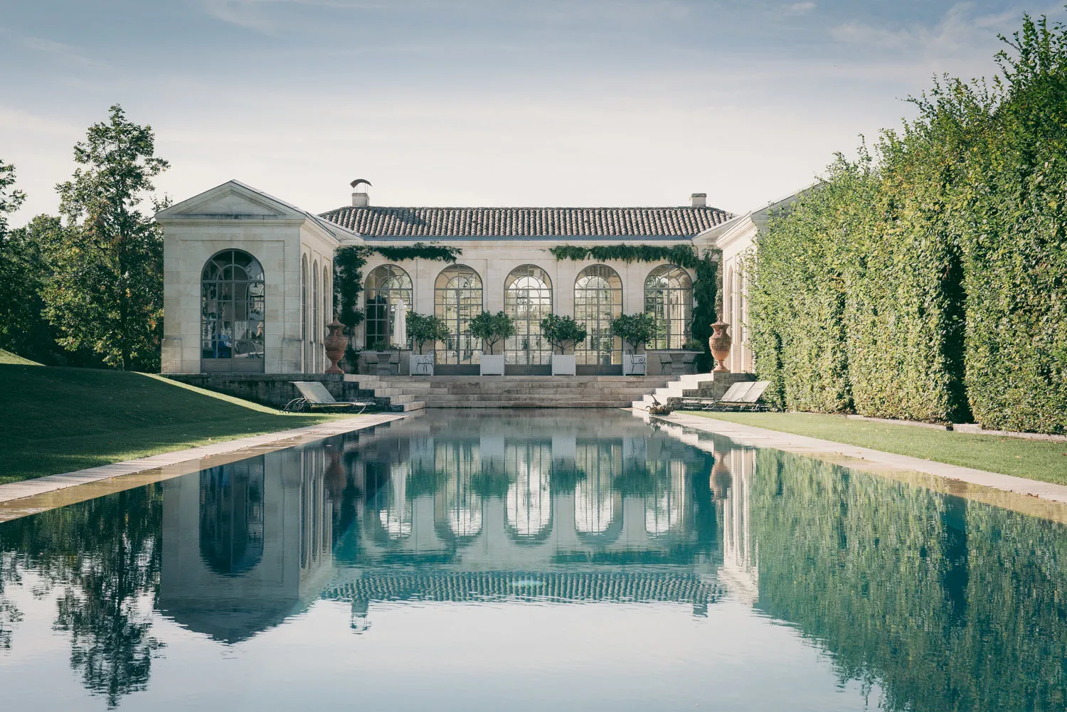 Bordeaux's Château du Tertre featuring its white neoclassical facade with arched windows, perfectly reflected in the rectangular pond, surrounded by cypress hedges and manicured gardens