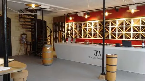 Modern wine cellar interior with DB reception desk, red walls, illuminated wooden wine racks, oak barrels and metal staircase at Château Lanessan in Bordeaux.
