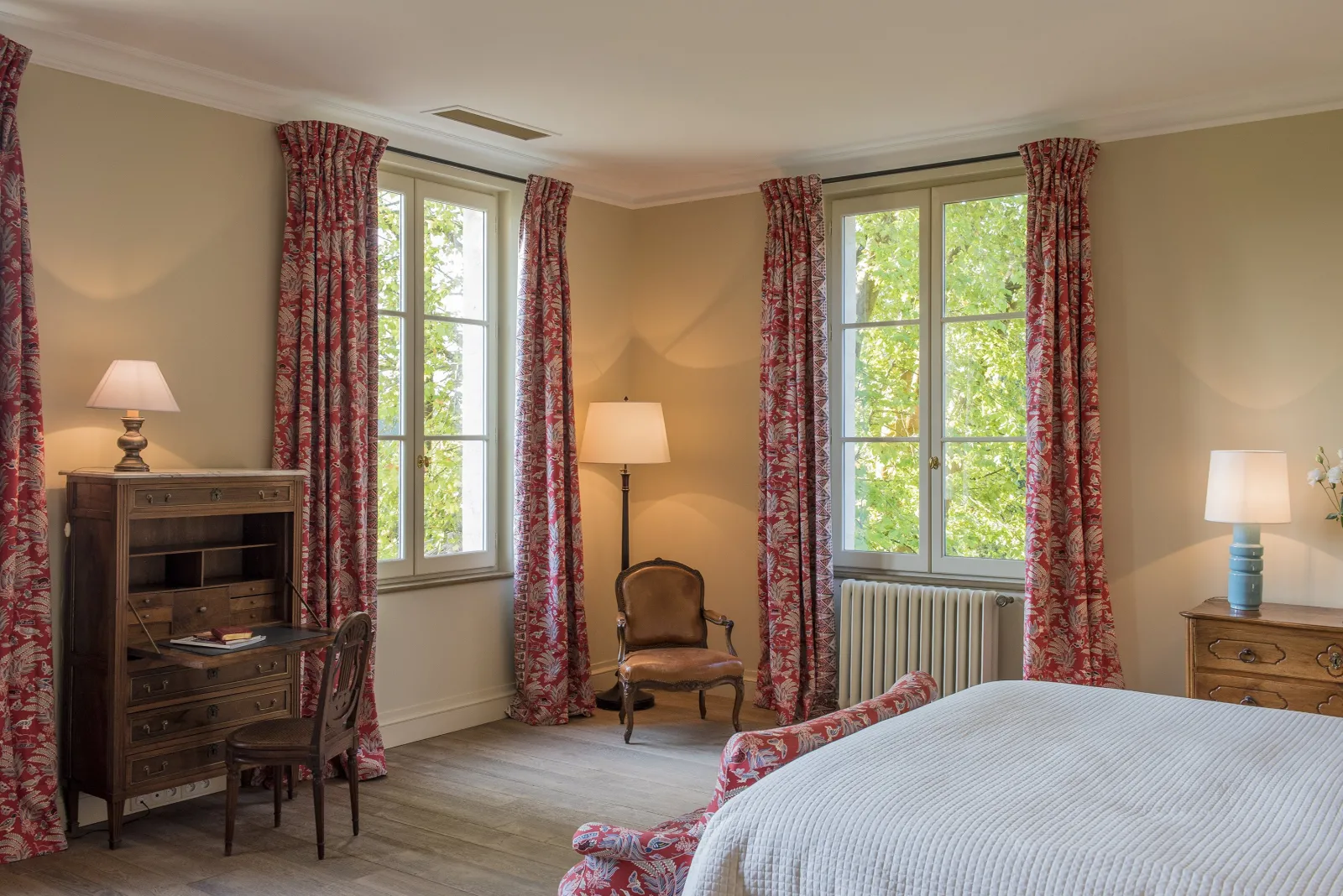 Elegant bedroom featuring a white bed, floral red and white patterned curtains, two French doors opening to a lush green garden, solid wood furniture, classic table lamps, and a brown armchair.