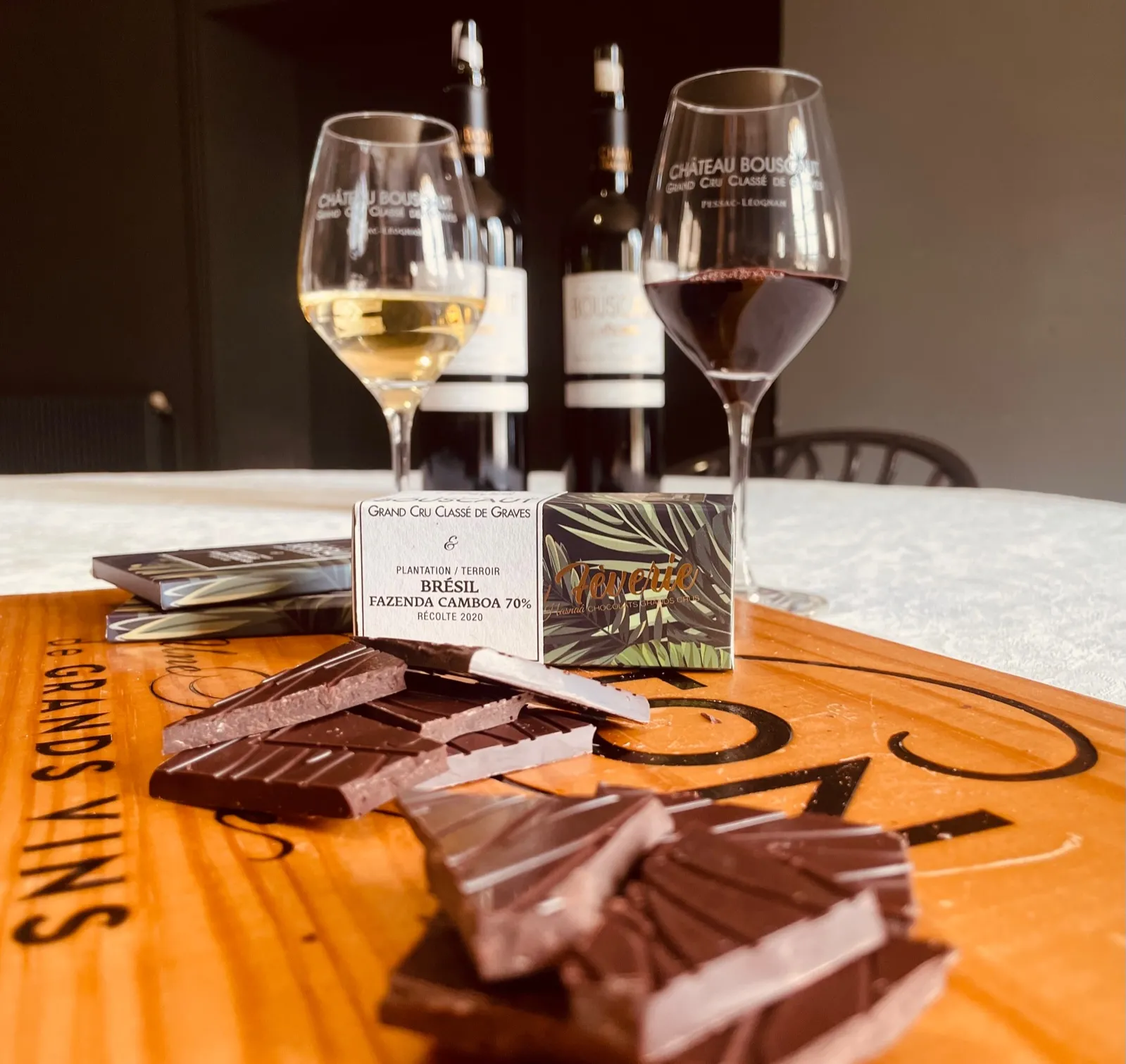 Wine and chocolate tasting at Château Bouscaut in Bordeaux: two glasses of white and red wine, chocolates arranged on an orange table, wine bottles in the background, and a Brazilian cocoa information card.