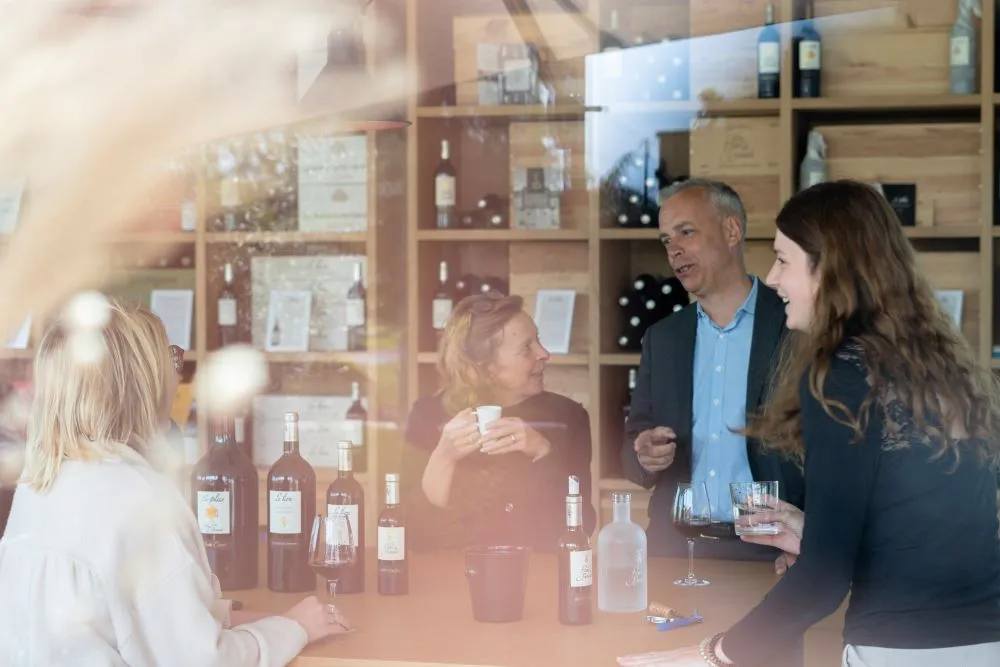 Wine tasting at Château La Fleur de Boüard in Bordeaux: four people conversing around a table with wine bottles and glasses, in front of wooden shelves displaying the estate's collection.