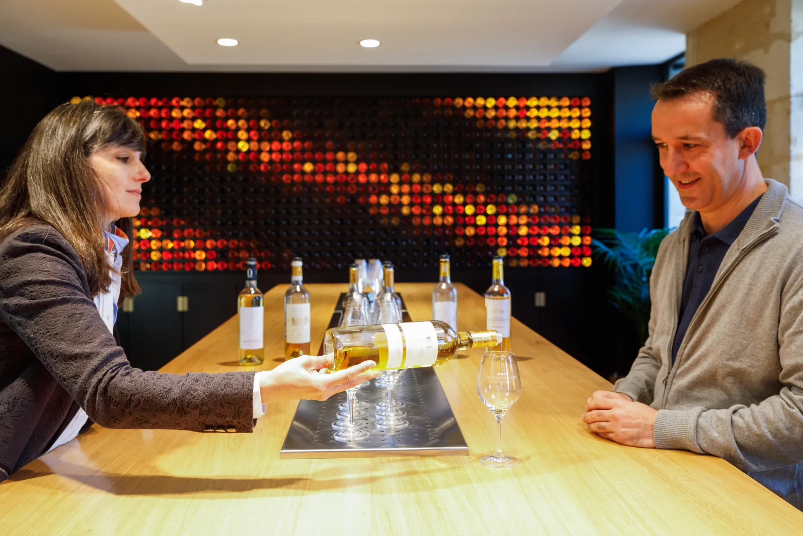 A woman pouring white wine into a glass while facing a smiling man in a tasting room with illuminated display and Sauternes bottles lined up on a wooden table.