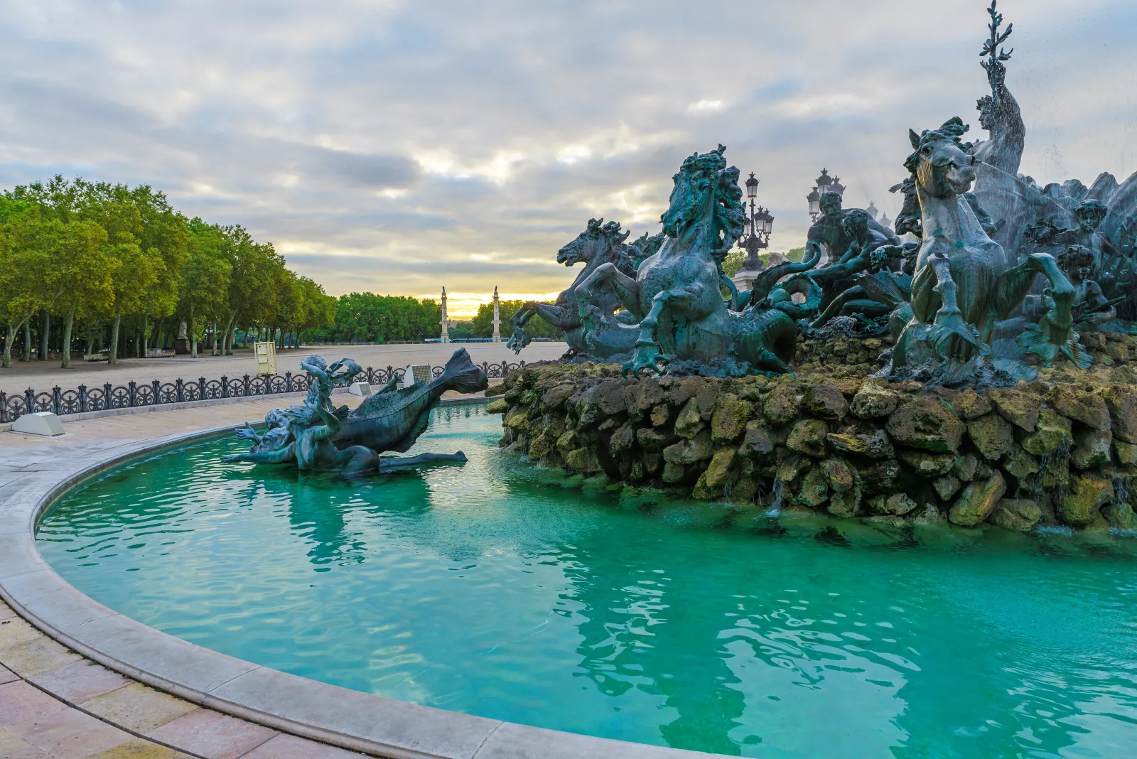 The Monument to the Girondins on the Quinconces Esplanade in Bordeaux, featuring green-patinated bronze statues overlooking a turquoise pool, surrounded by trees and ornamental railings.