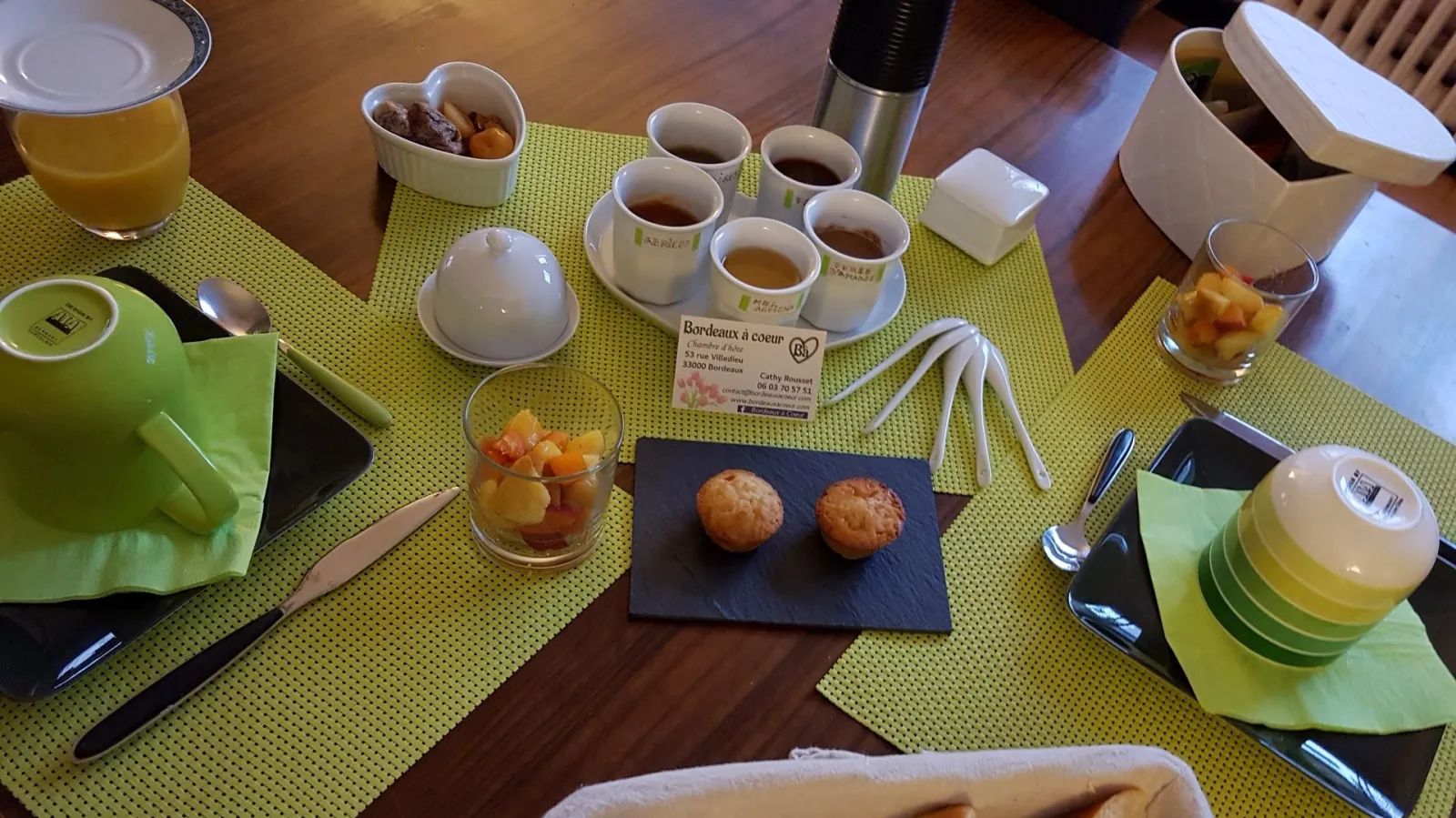 Breakfast table with lime-green placemat, orange juice and fruit juice glasses, coffee cups, pastries, fresh fruit, and a Bordeaux à Cœur business card