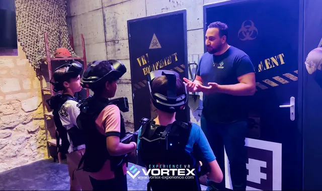 An instructor briefs a group of visitors wearing virtual reality headsets on safety instructions at a VR experience zone in Bordeaux, with blue doors marked with hazard symbols in the background.