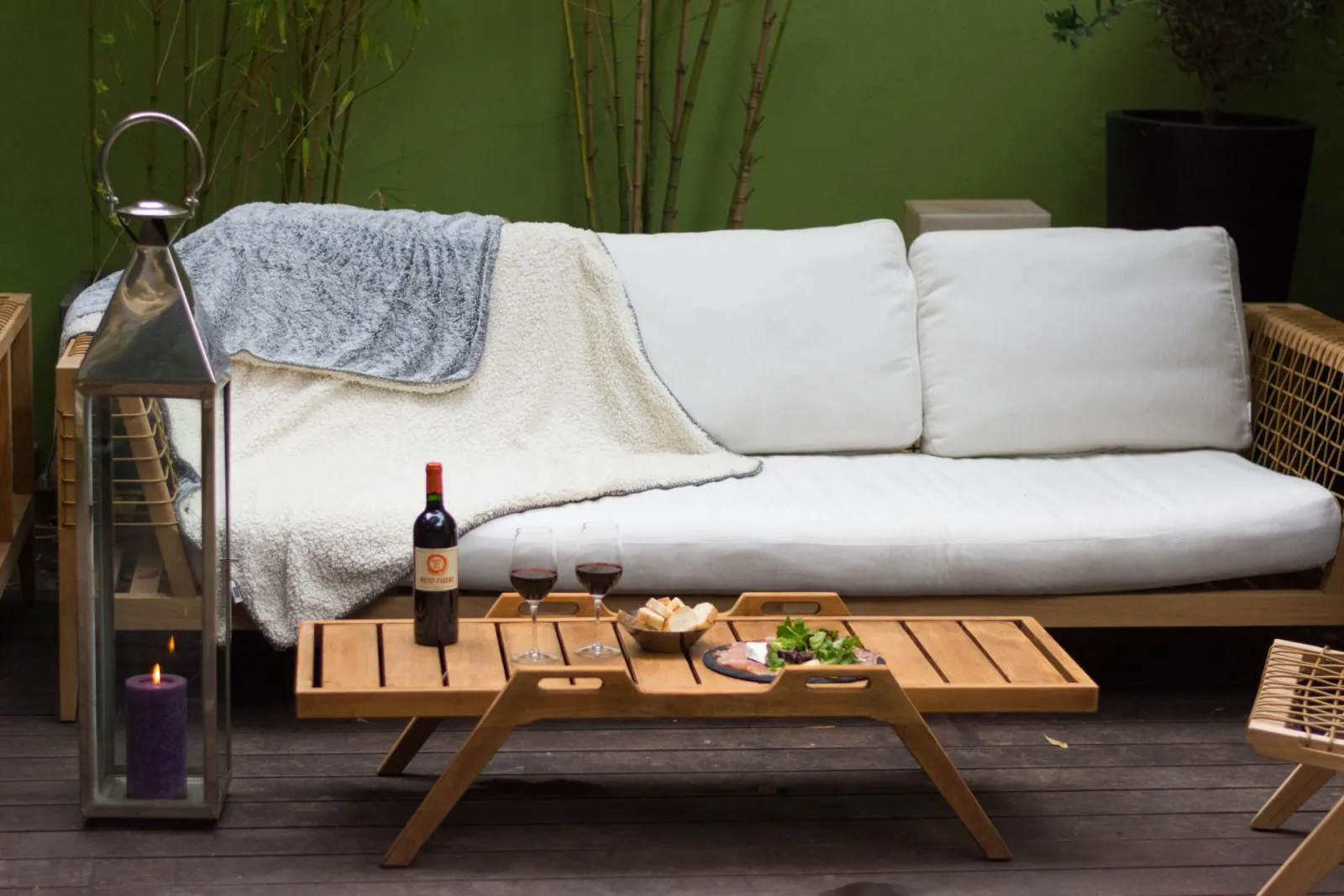 Elegant outdoor terrace featuring a white sofa, wooden coffee table with Bordeaux wine bottle, glasses, charcuterie and salad, black lantern with purple candle, grey and beige throws, bamboo plants in background, relaxing atmosphere