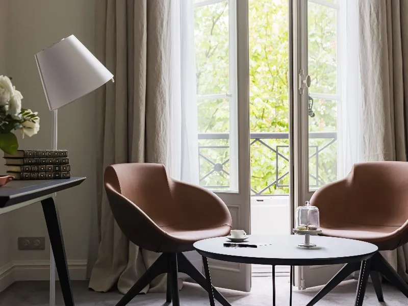 Elegant living room with two dusty pink armchairs, round white coffee table, desk with lamp, and large glass doors opening onto a balcony overlooking greenery at Hôtel Villas Foch in Bordeaux.