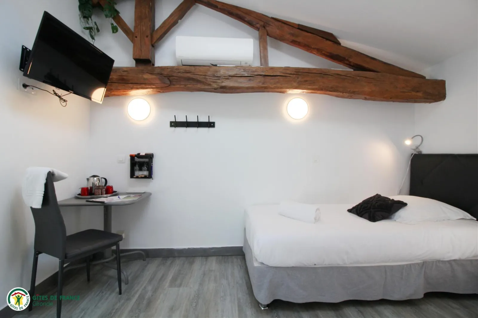 Modern guest room featuring a queen-size bed, black desk chair, exposed wooden beams, contemporary lighting, and wall-mounted TV at Château Pierre de Montignac in Bordeaux.