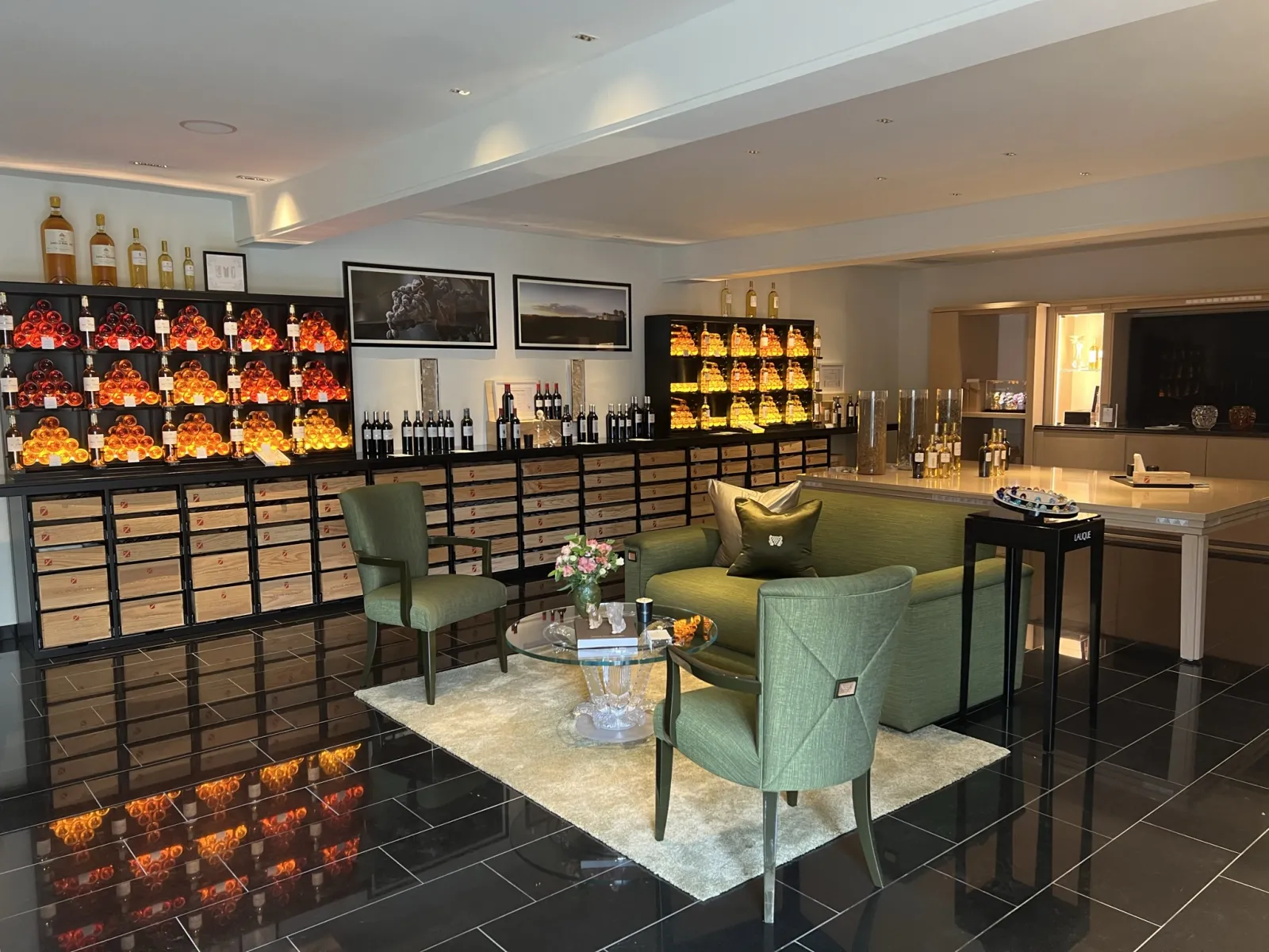 Elegant tasting room at Château Lafaurie Peyraguey in Bordeaux, featuring illuminated wine bottle displays, green armchairs, bar counter, and glass wine cellar.