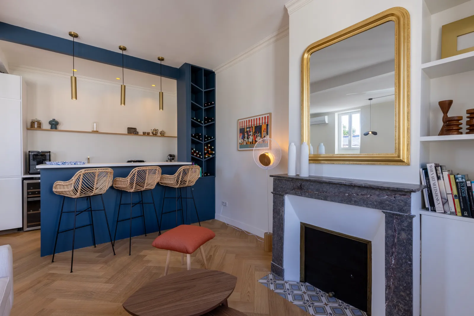 Modern apartment interior in Bordeaux featuring a navy blue open kitchen, bar with three rattan chairs, black marble fireplace, gold mirror, white shelving and herringbone parquet flooring.
