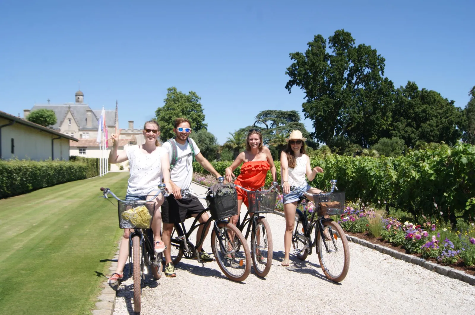 Four smiling cyclists pose with their bikes on a gravel path in Bordeaux vineyards, with a château and trees in the background.