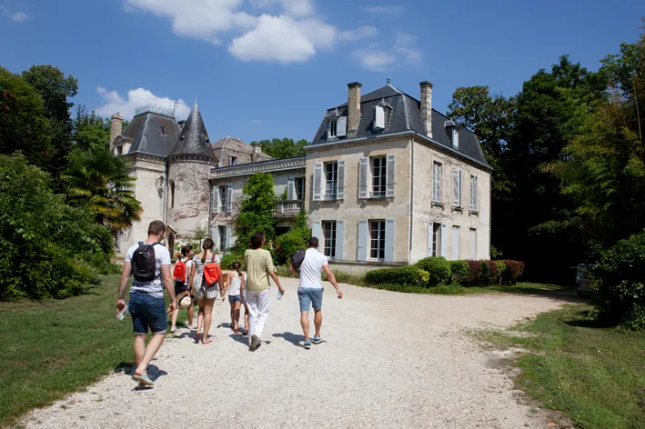 Group of visitors walking on a gravel path in front of Château Bardins, a 19th-century mansion with turrets and mansard roofs, surrounded by greenery near Bordeaux.