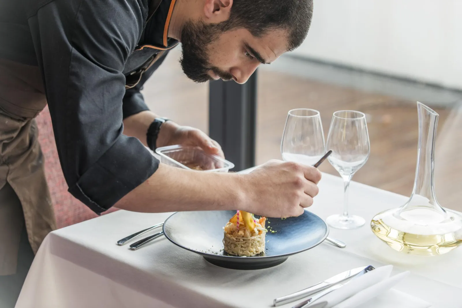 A professional chef in chef's attire carefully plating a gourmet dish on a blue plate, with wine glasses and a decanter visible in an upscale restaurant setting.
