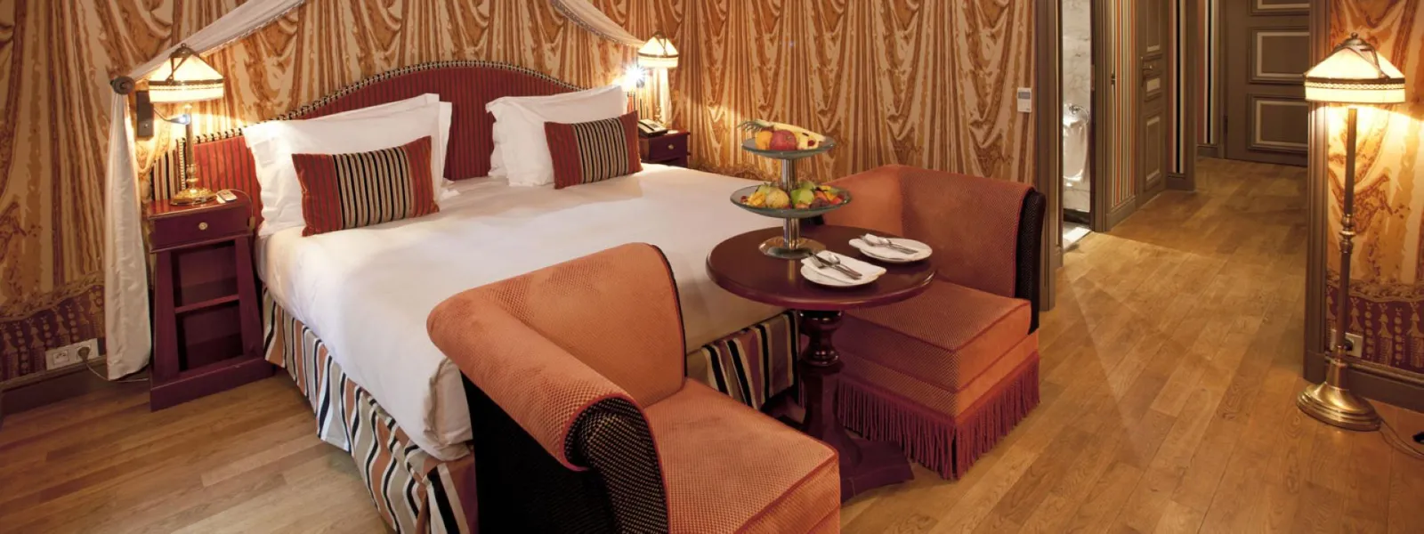 Luxury suite at the Intercontinental Grand Hotel Bordeaux featuring a white and red king-size bed, mahogany furniture, golden drapes, brass floor lamps, and a seating area with side table and fruit platters