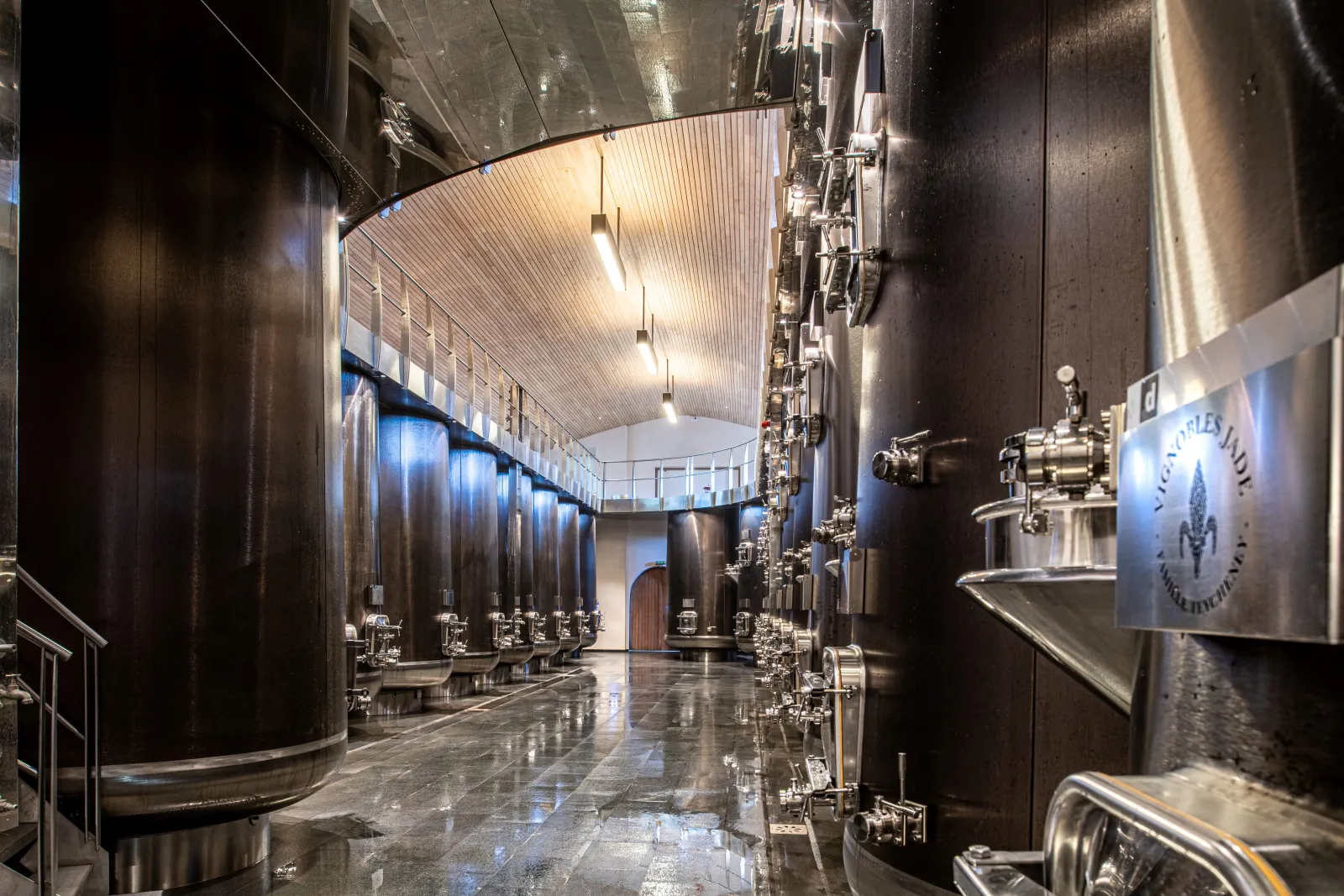 Interior of a winery cellar featuring large stainless steel fermentation tanks lined on both sides of a corridor, equipped with control valves, under a metal beam ceiling with pendant lighting.