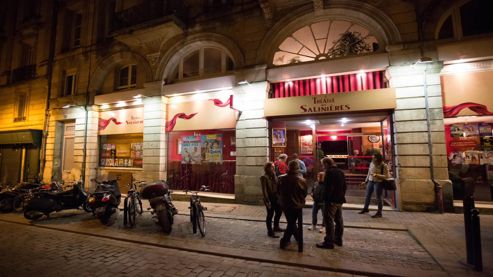 Night view of the Théâtre des Salinières in Bordeaux, featuring its distinctive red entrance, parked bicycles and scooters, and pedestrians on the cobblestone plaza.