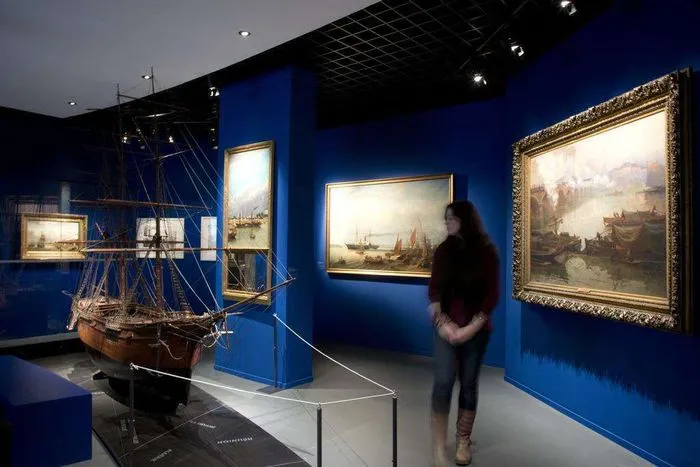 Museum gallery with deep blue walls displaying a wooden ship model, framed maritime paintings with golden frames, and a visitor viewing the historical exhibits