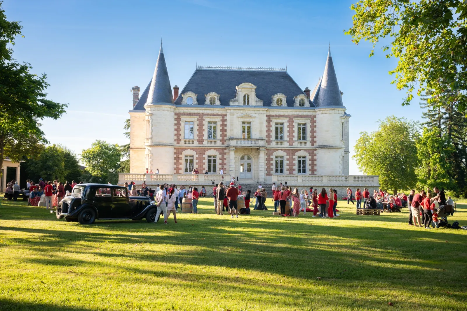 Château Lamothe Bergeron in Bordeaux, a grand neo-Renaissance castle with white stone and red brick turrets, hosting an outdoor reception with numerous guests dressed in red on the lawn under clear skies.