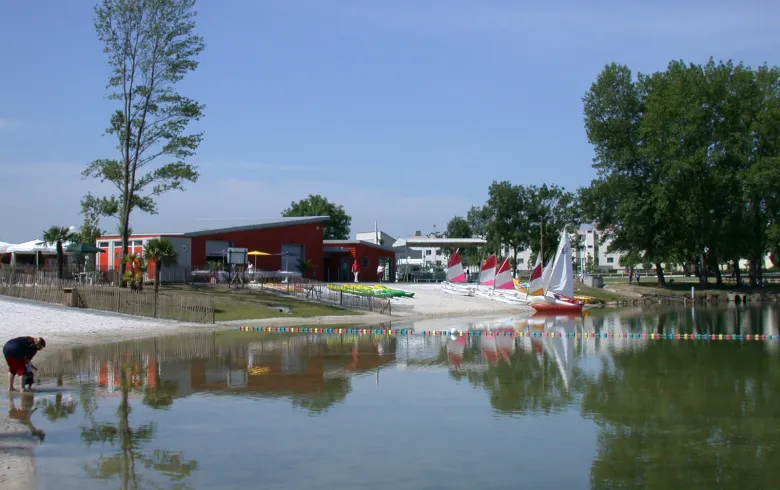 Bègles Beach in Bordeaux featuring a red building by the water, colorful sailboats, trees, and a person in the foreground
