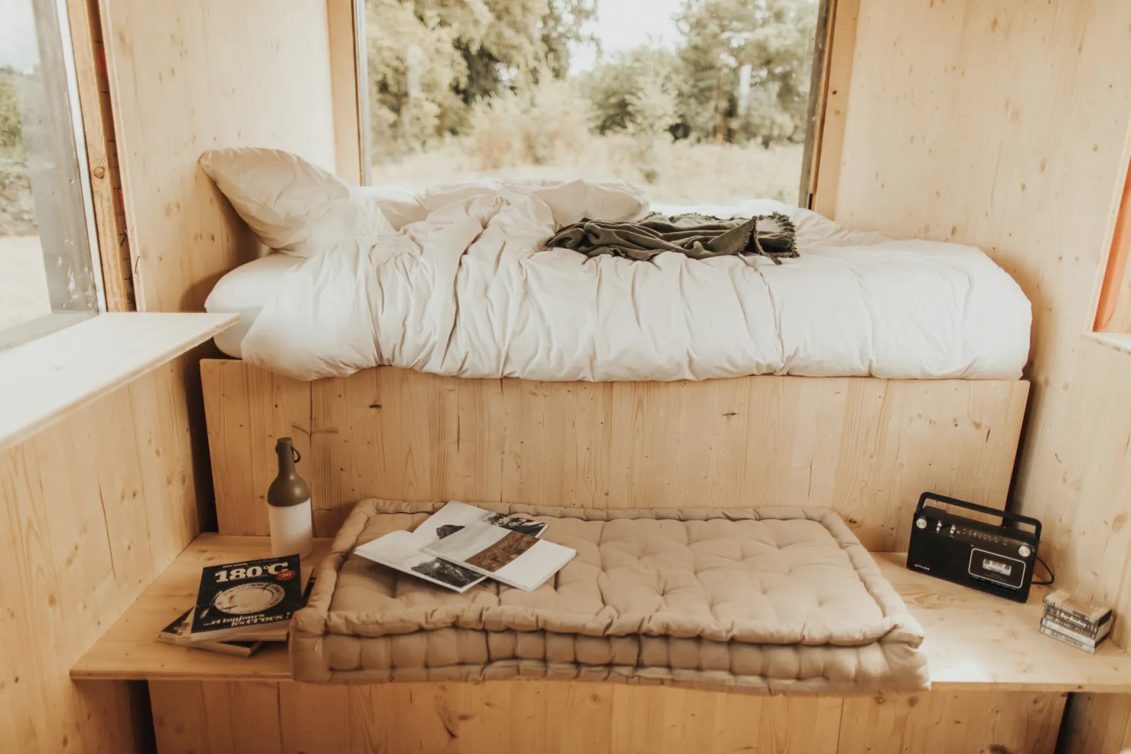 Minimalist wooden tiny house interior featuring a bunk bed with beige bedding, a futon on the lower level, large windows overlooking forest scenery, and vintage décor.
