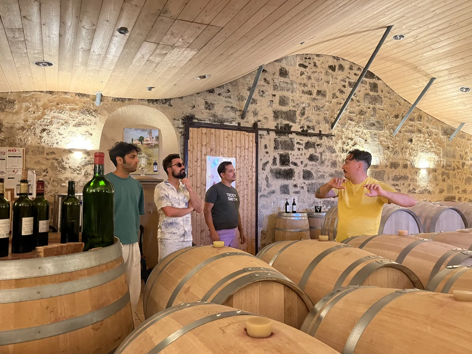 Guided tour of a Bordeaux wine cellar featuring oak barrels and four visitors listening to the winemaker's explanation in an underground stone-walled cellar.