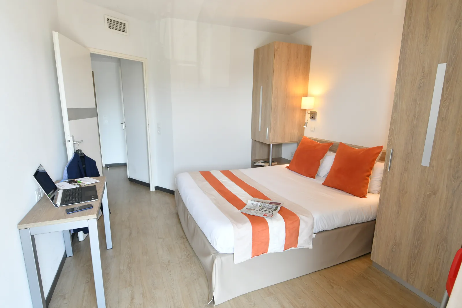 Modern hotel apartment room featuring a double bed with orange and white striped bedding, work desk with laptop, light wood wardrobe, ensuite bathroom, and minimalist décor.